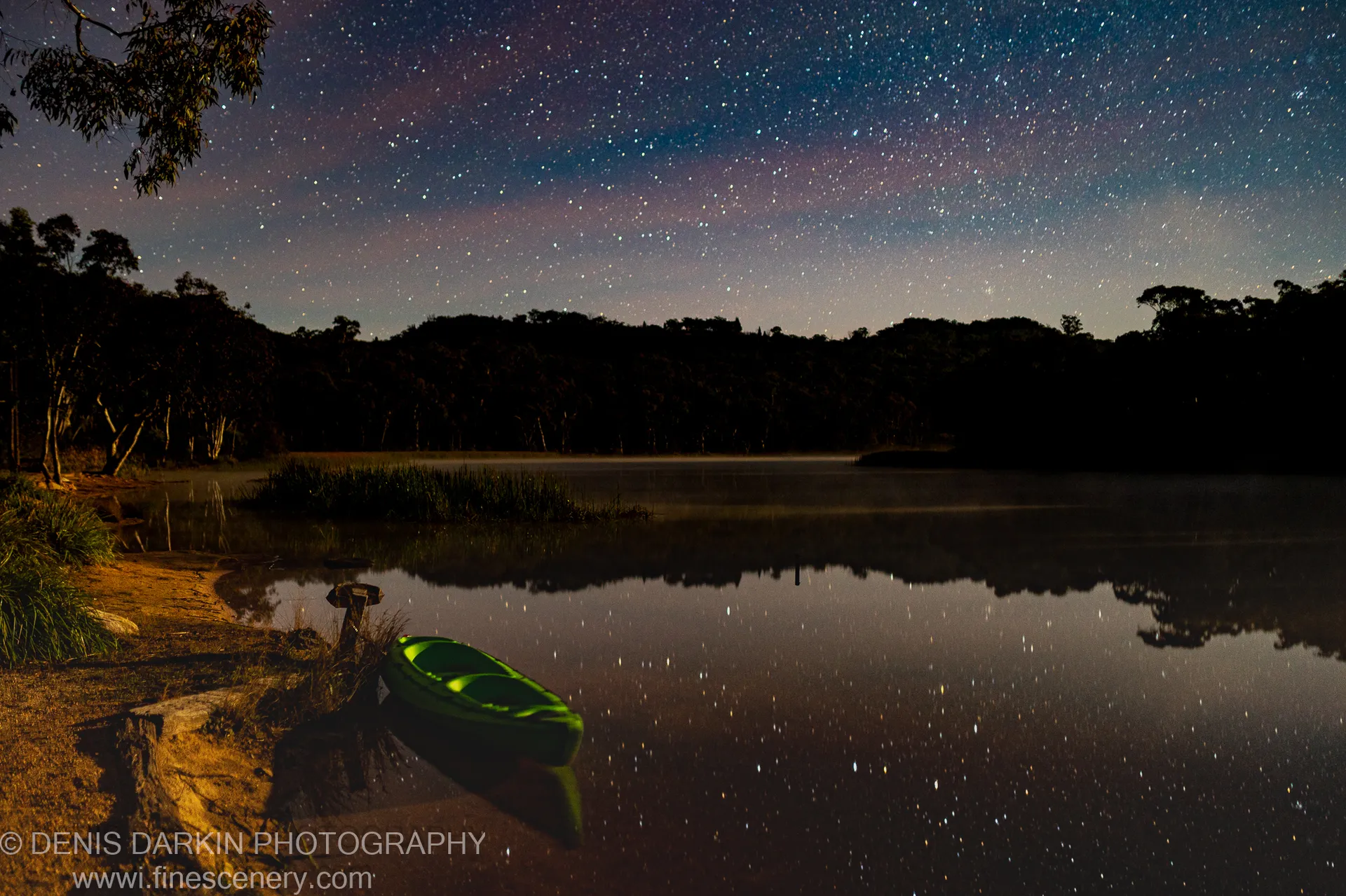 A bright green kayak rests on the bank of Dunns Swamp while a starfield and its near-perfect reflection fill the frame above and below.