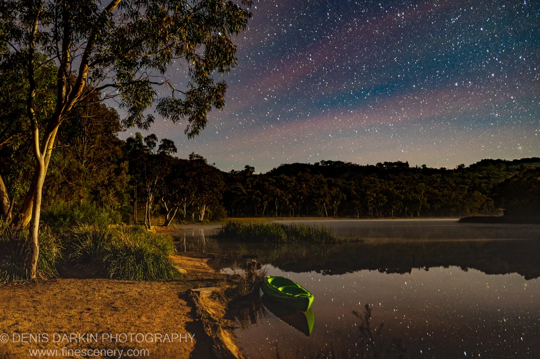 A warm-lit green canoe rests at a sandy bank at Dunns Swamp at night, stars reflected in the glassy water around it.