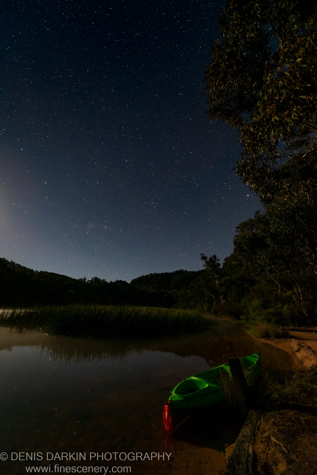 A bright green kayak sits at a sandy shore on Dunns Swamp at night, dark silhouetted hills rising beneath a dense starfield.