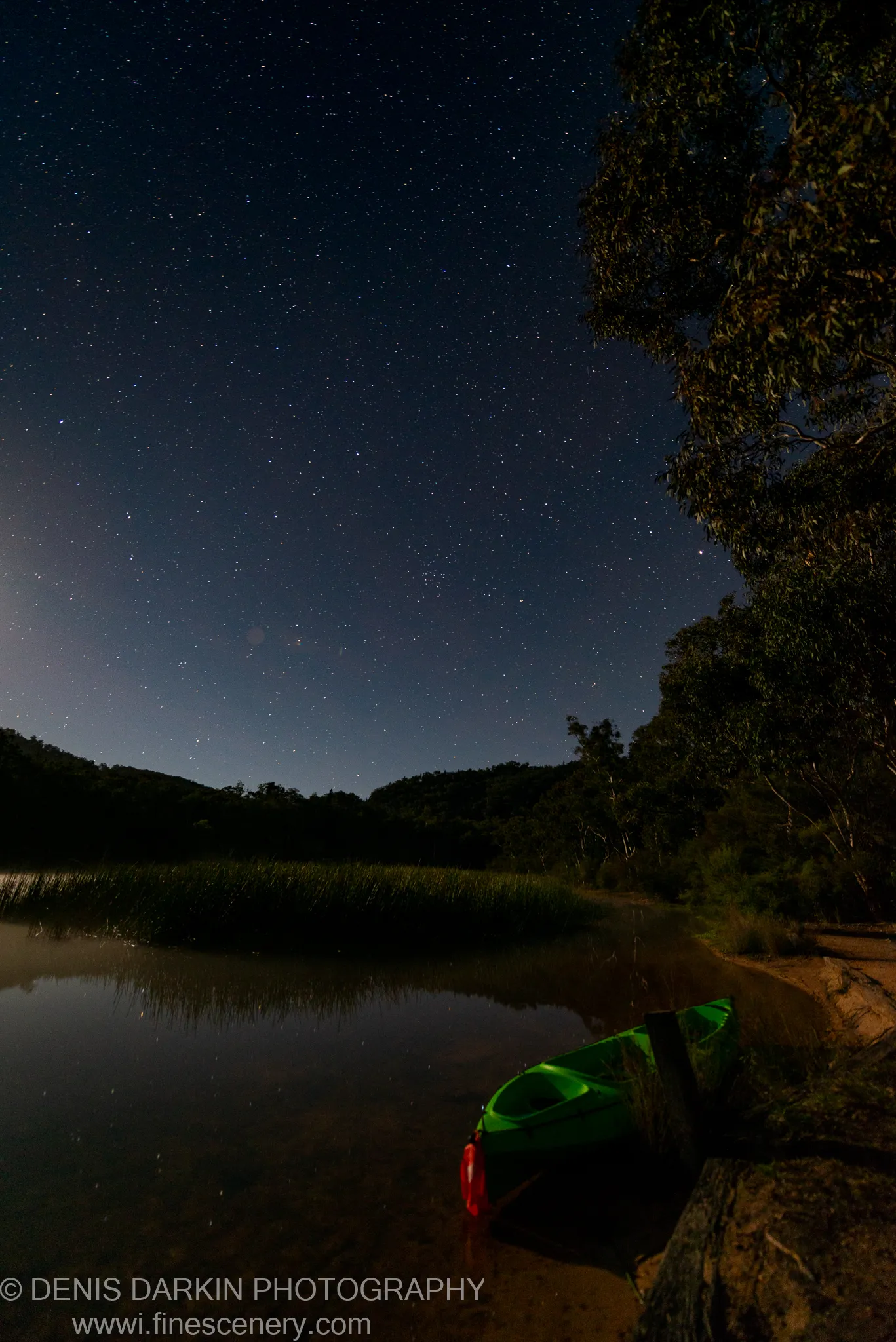 A bright green kayak sits at a sandy shore on Dunns Swamp at night, dark silhouetted hills rising beneath a dense starfield.