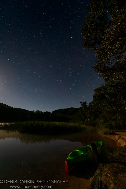 A bright green kayak sits at a sandy shore on Dunns Swamp at night, dark silhouetted hills rising beneath a dense starfield.