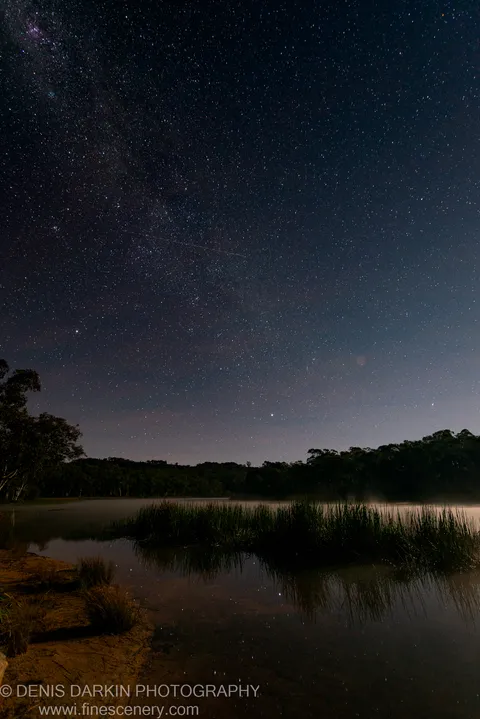 Dunns Swamp at night with a faint Milky Way arc visible above still water that mirrors the stars and reed silhouettes below.