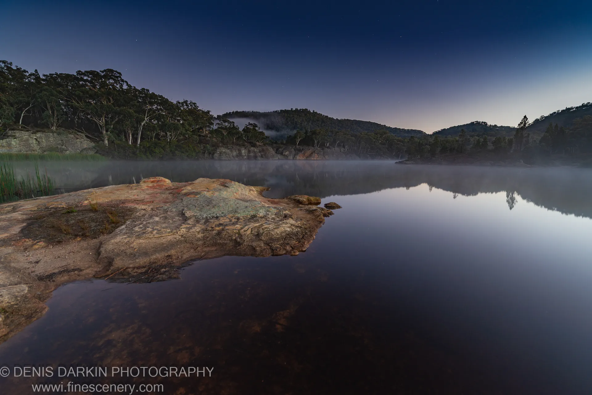 A lichen-covered rock formation sits beside the glassy water of Dunns Swamp during blue hour, low mist clinging to the still surface.