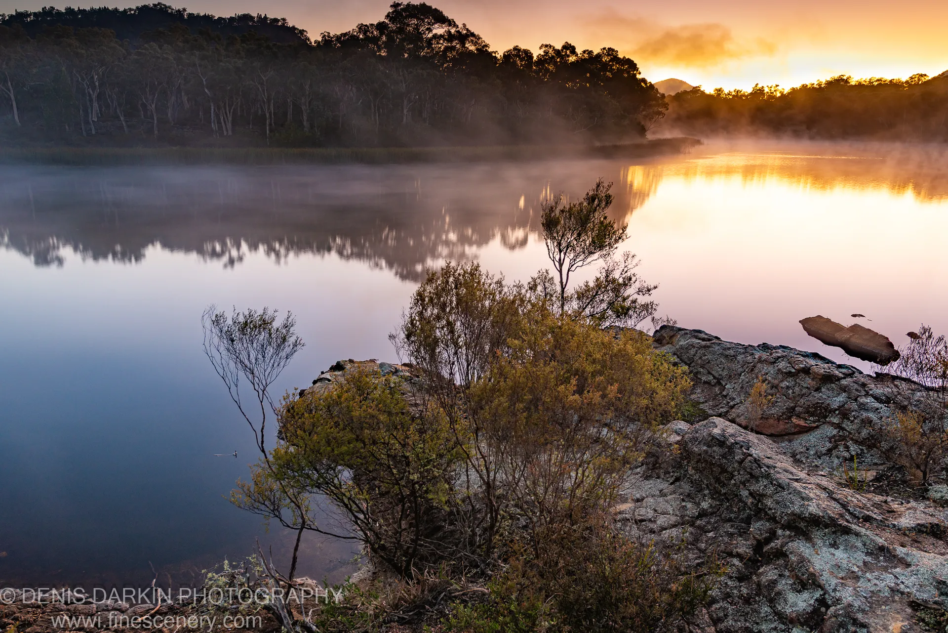 Lichen-covered rocks in the foreground with mist over still water and warm dawn reflections at Dunns Swamp, New South Wales.