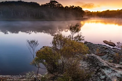 Lichen-covered rocks in the foreground with mist over still water and warm dawn reflections at Dunns Swamp, New South Wales.