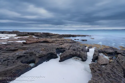 Pockmarked sandstone rock shelf with foam-filled pools at Huskisson, New South Wales, under an overcast predawn sky in December 2019.