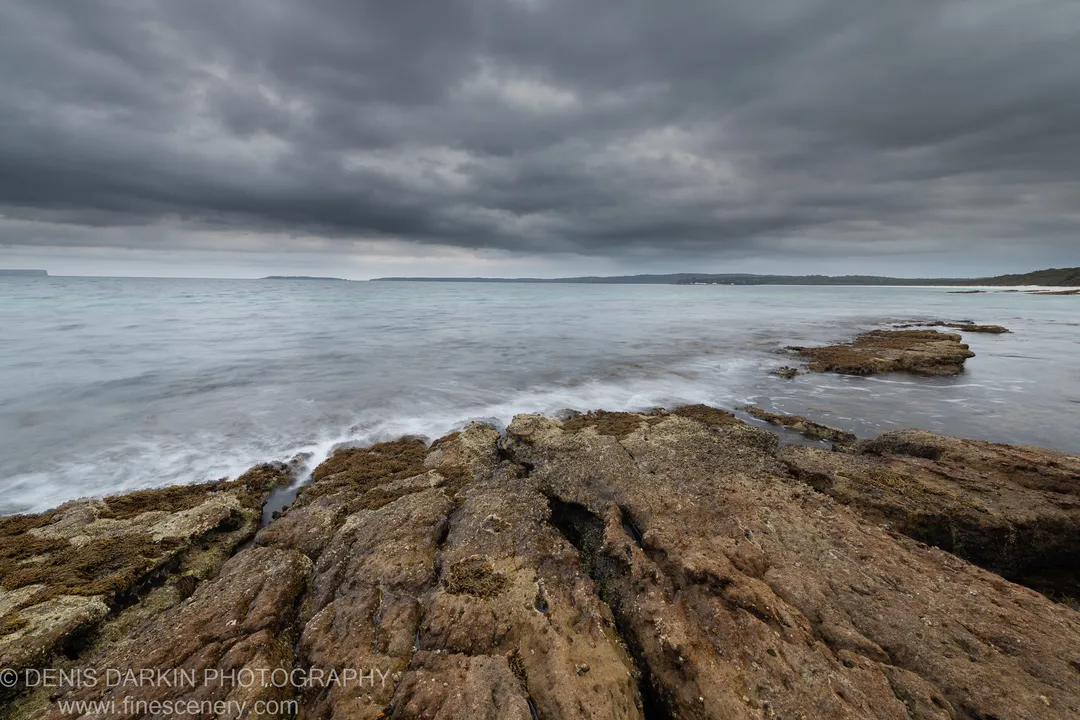 Long-exposure seascape with layered rock platform and pale milky water under dark storm clouds at Huskisson, New South Wales, December 2019.
