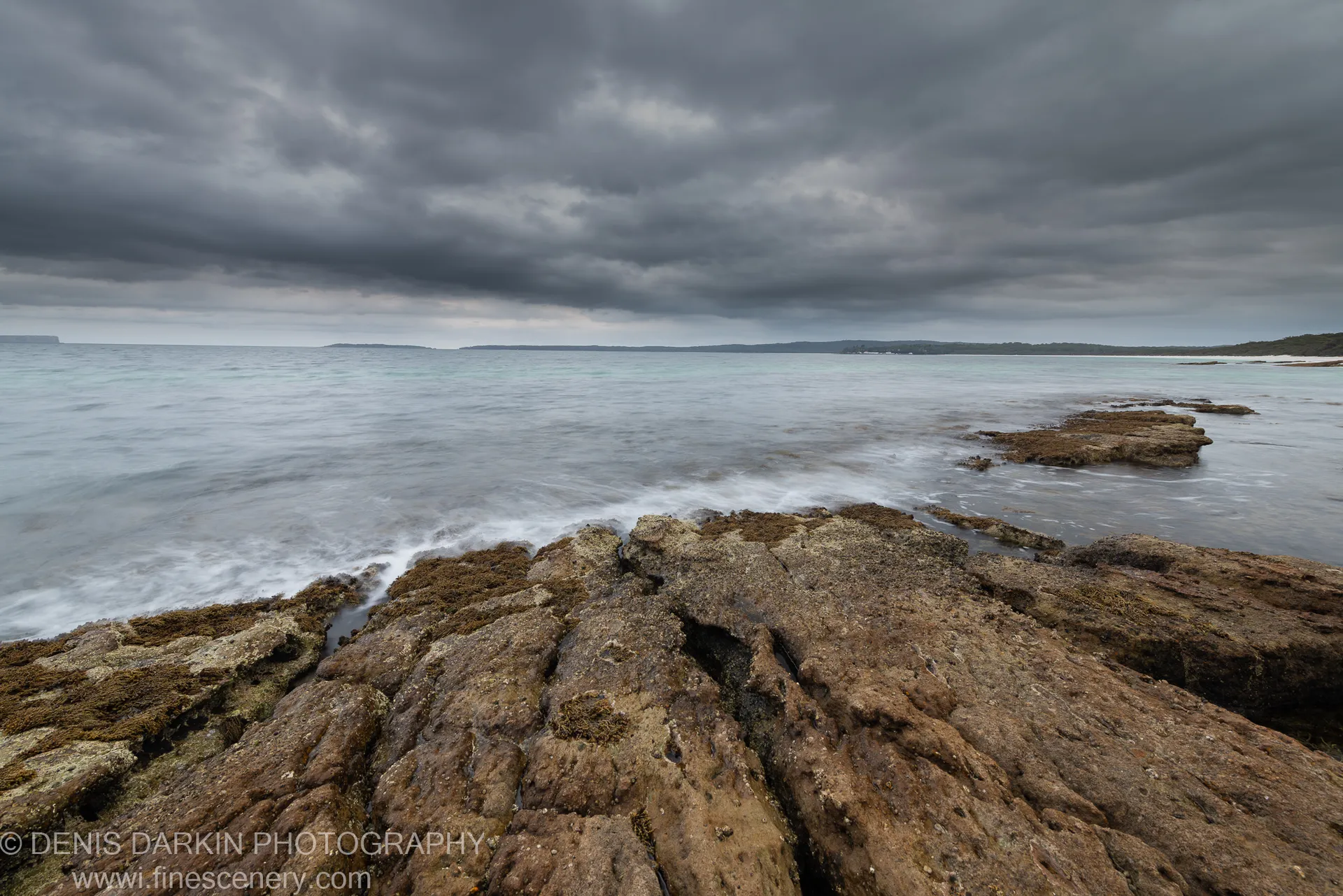 Long-exposure seascape with layered rock platform and pale milky water under dark storm clouds at Huskisson, New South Wales, December 2019.