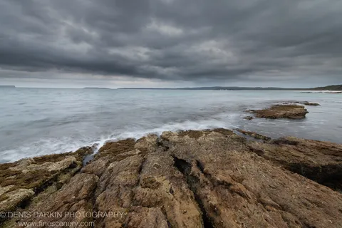Long-exposure seascape with layered rock platform and pale milky water under dark storm clouds at Huskisson, New South Wales, December 2019.