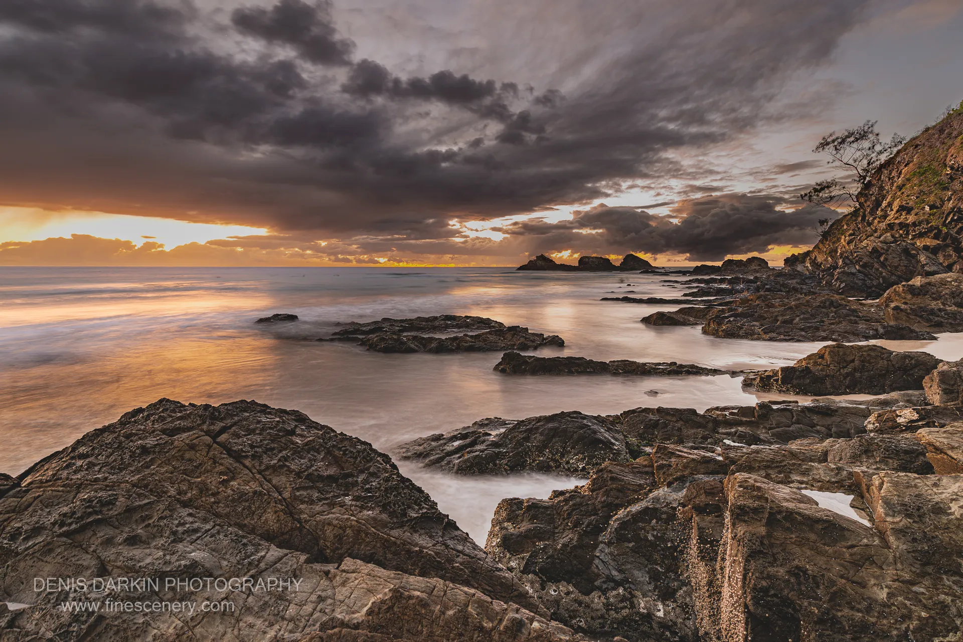 Volcanic rocks in the foreground with golden light breaking through storm clouds over long-exposure ocean surf at Byron Bay on a winter dawn.