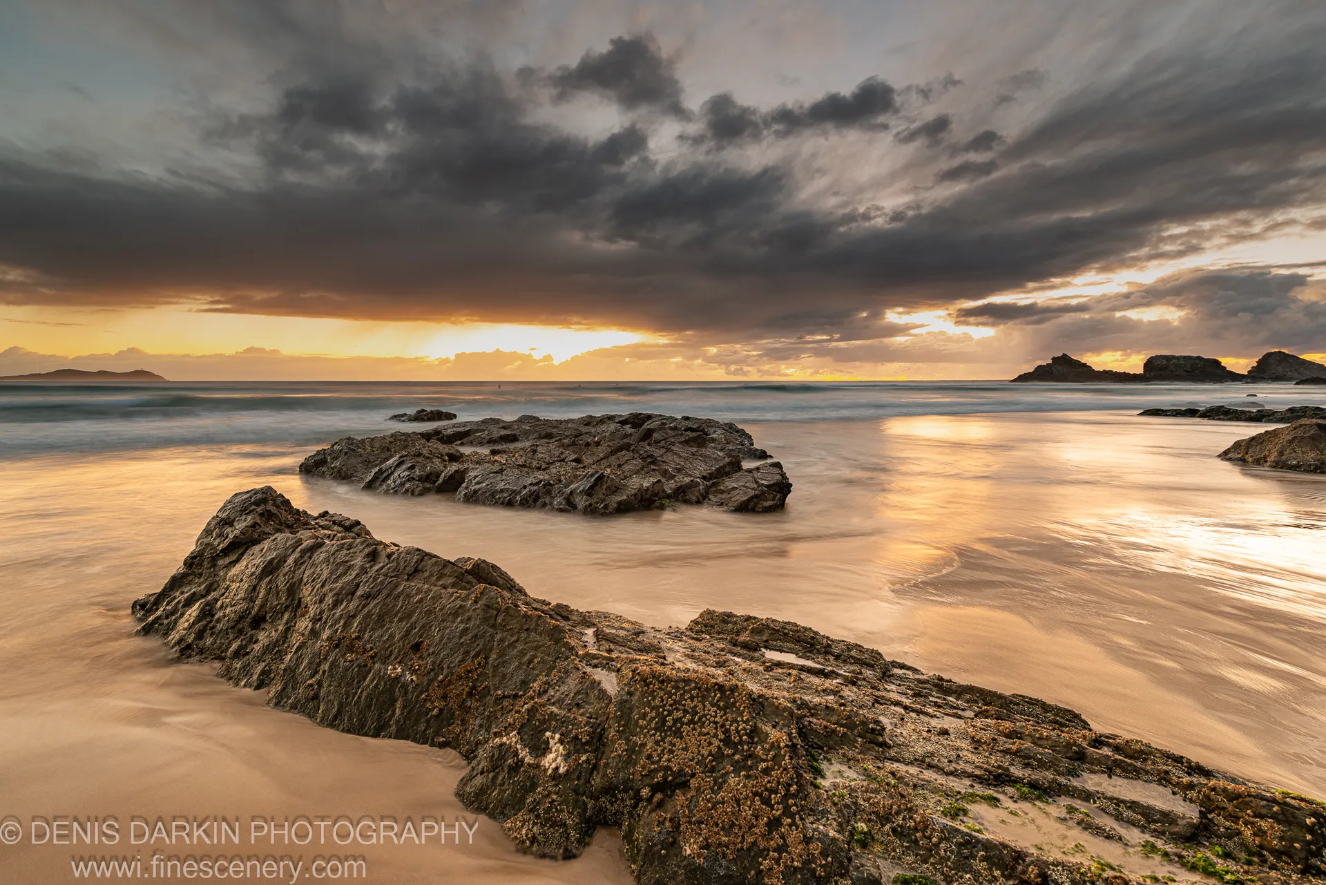 Barnacle-covered rocks in the foreground of a Byron Bay coastal scene, golden light breaking through storm clouds over smooth water.