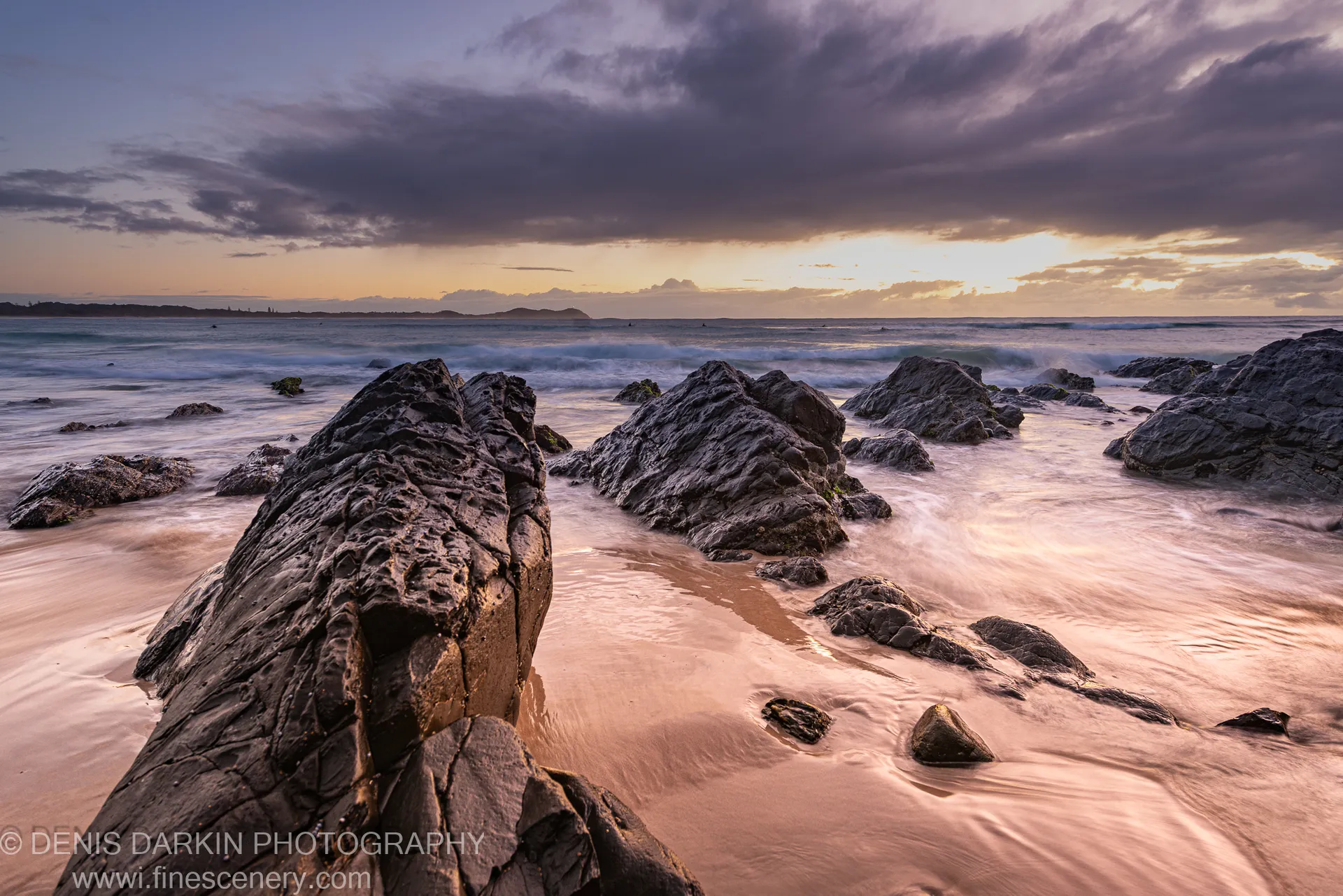 Dark rock formations meet long-exposure surf at Byron Bay dawn, golden light washing over wet sand beneath a purple-tinged winter sky.