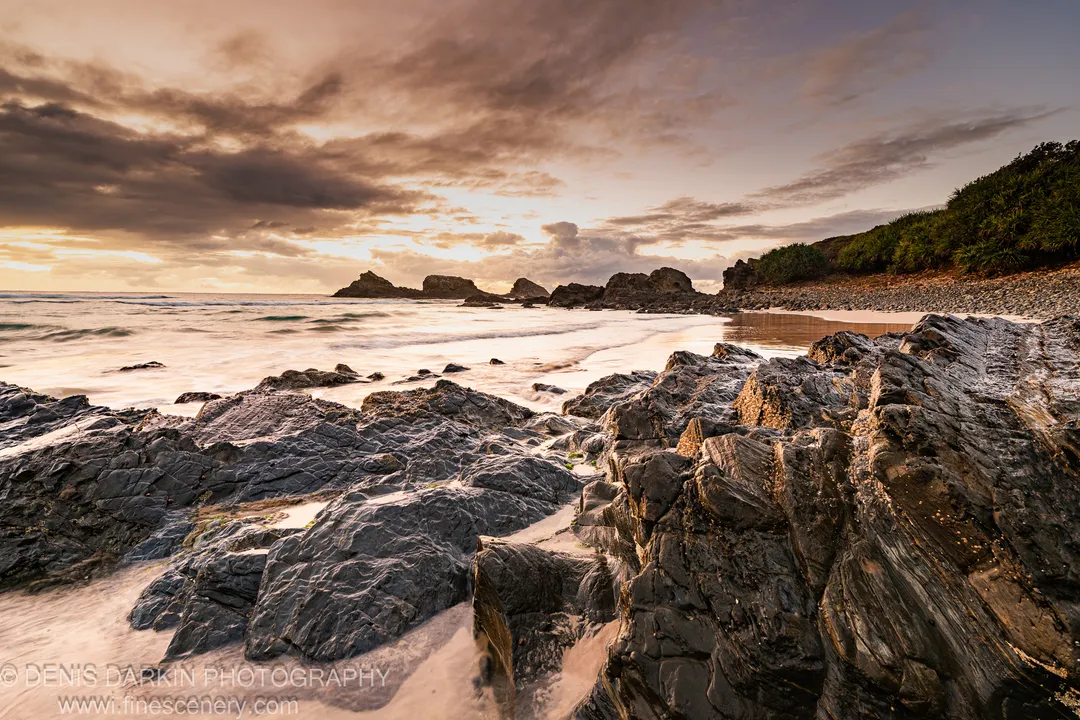 Stratified coastal rocks in the foreground at Byron Bay dawn, golden-lit clouds above the headland and sea stacks visible in the distance.