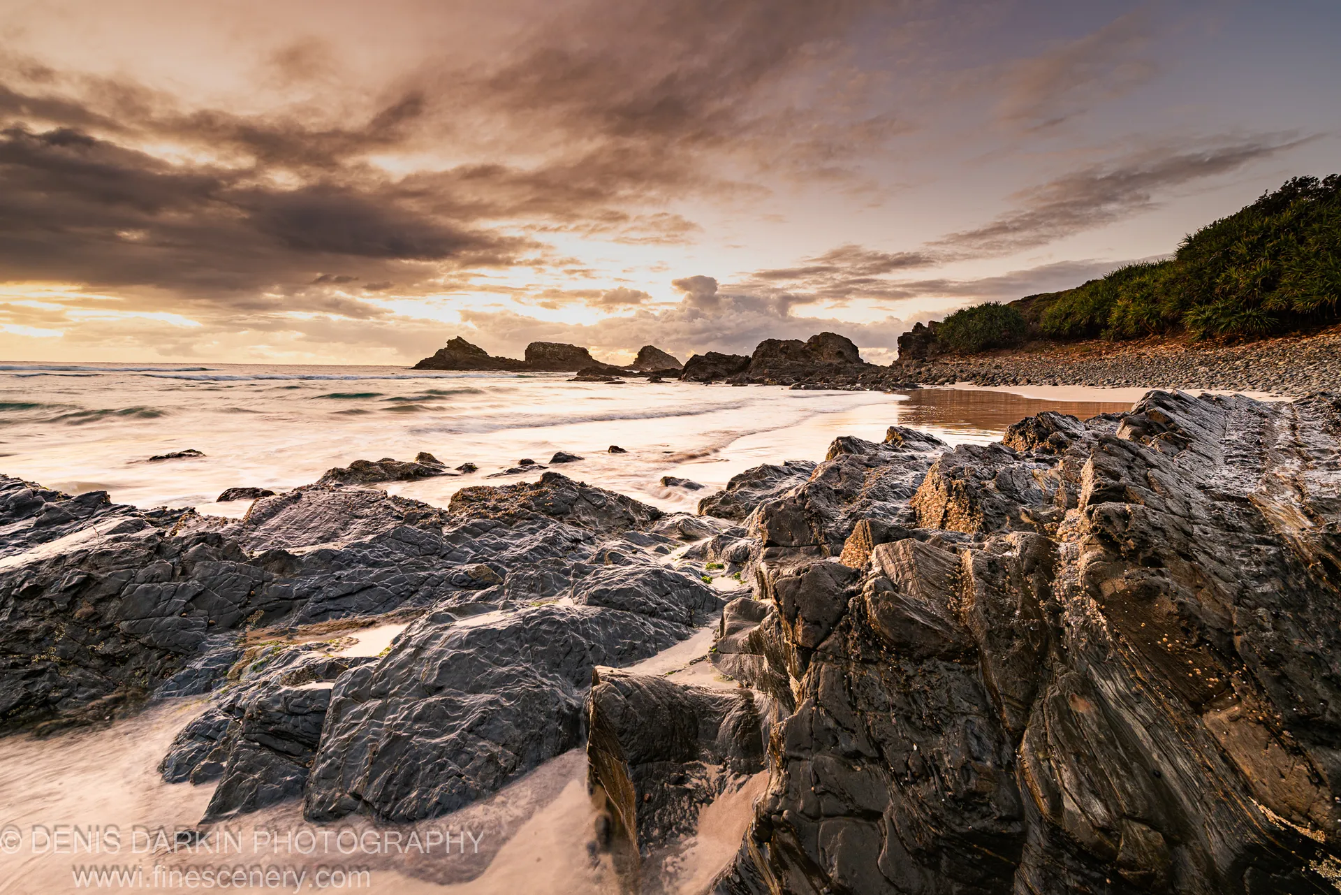 Stratified coastal rocks in the foreground at Byron Bay dawn, golden-lit clouds above the headland and sea stacks visible in the distance.