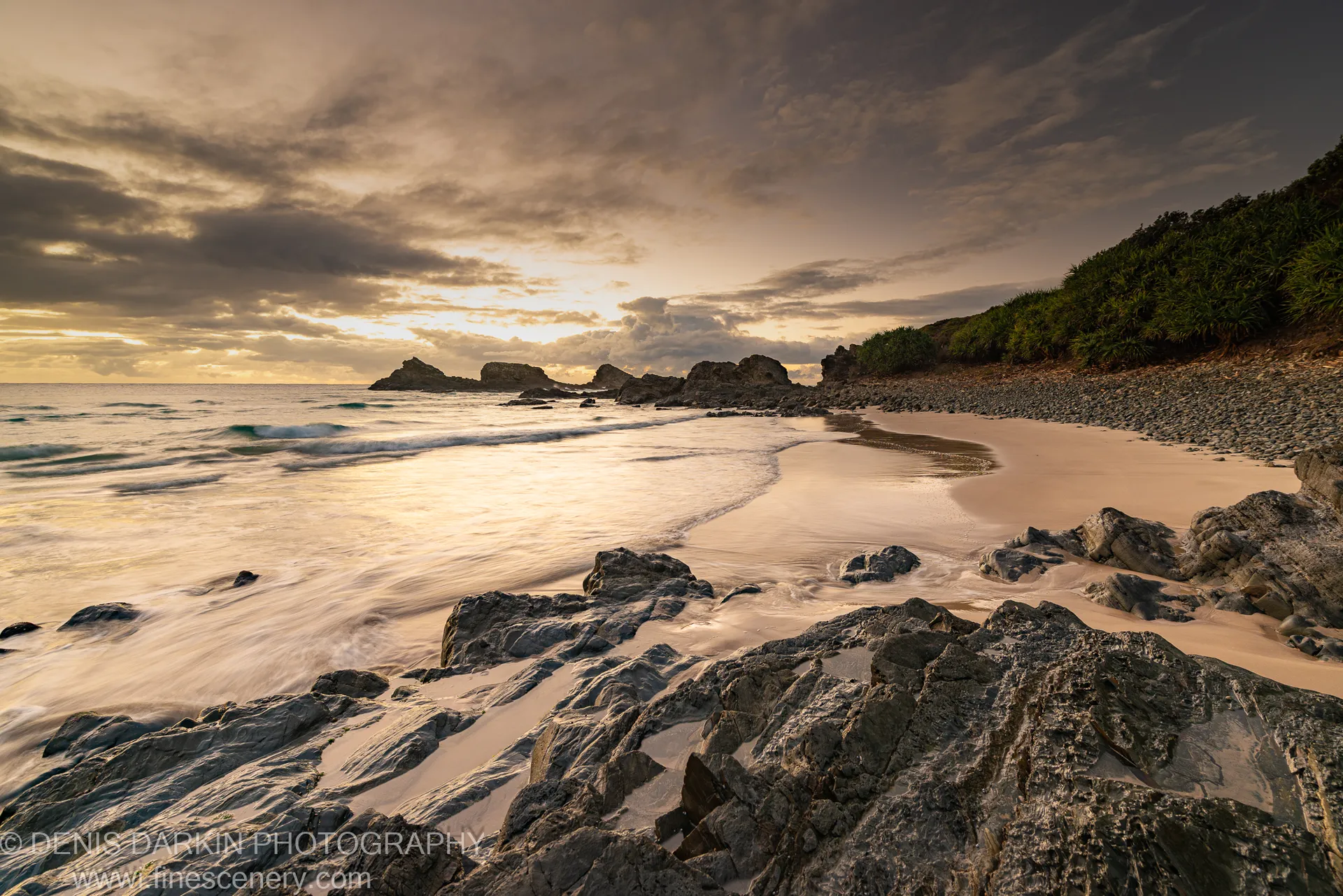 Grey boulders lead toward a sea stack silhouetted against clouded sky at Byron Bay, long-exposure surf forming a pale wash below.