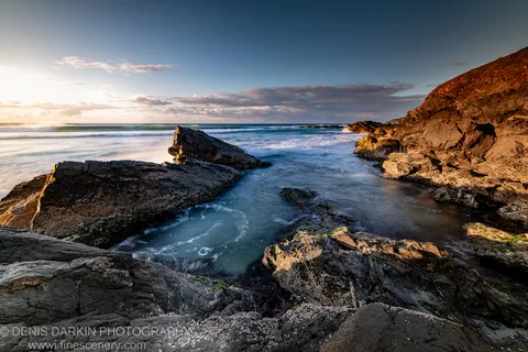 Barnacle-crusted rocks at a Byron Bay tidal channel with golden light, the ocean's flow smoothed into a pale stream against cool blue water.