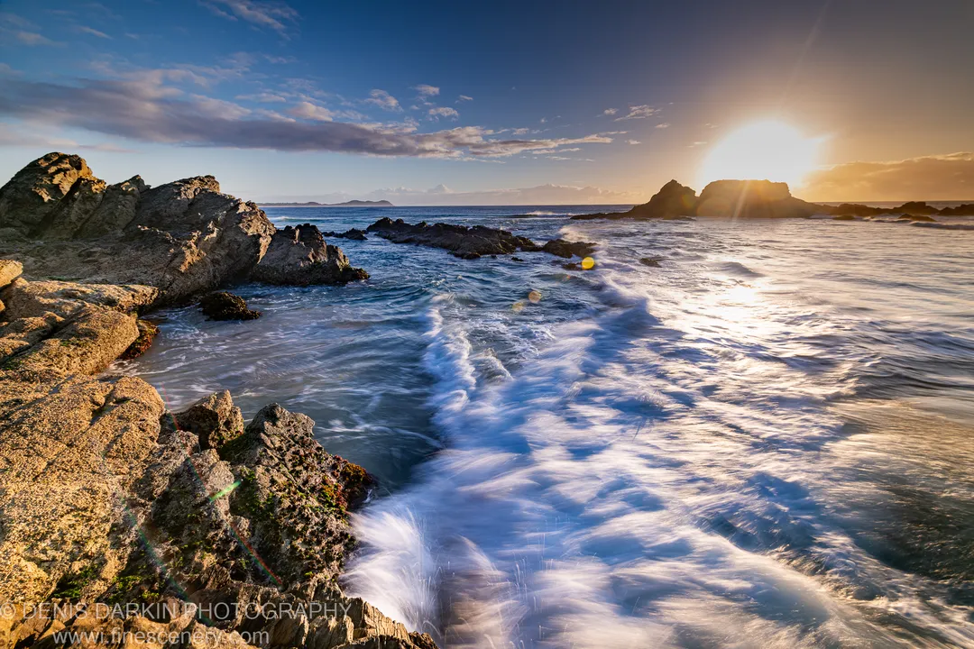 Starburst lens flare above a Byron Bay coastal channel at dawn, white surf rushing past mossy granite formations in long exposure.