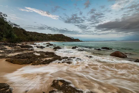 Rugged Queensland coastline at dawn, long-exposure waves softened to pale streaks across dark rocks below a curving tree-lined headland.