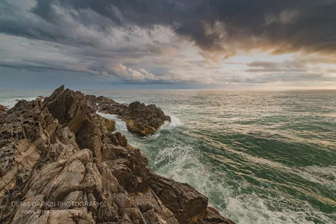 Fractured rock formations extend into the ocean on the Queensland coast, dark storm clouds above and a strip of warm light cutting across the horizon.