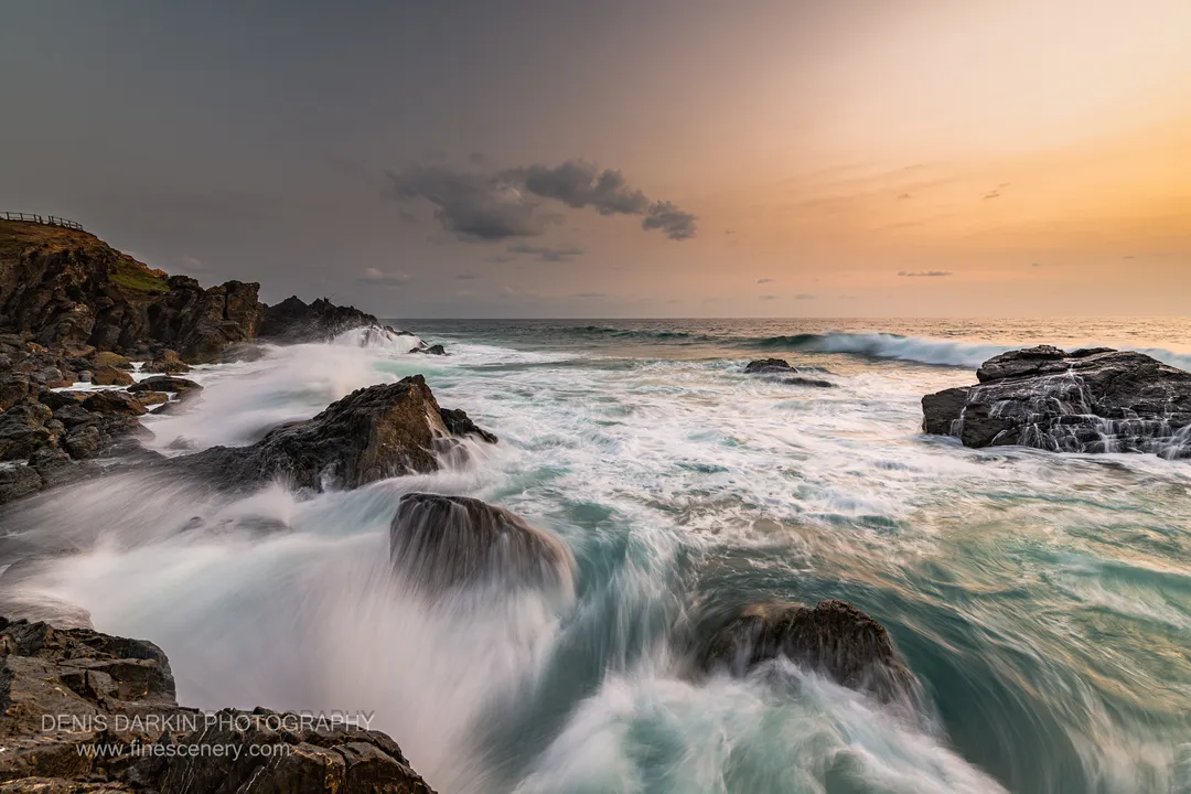 Jagged rocks rise in the foreground as long-exposure waves roll across the Queensland coast beneath a split sky of cool grey and warm gold.
