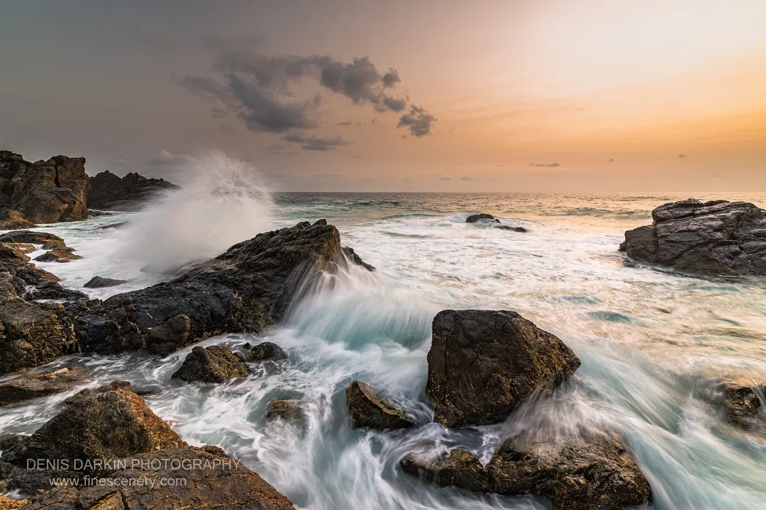 Long-exposure waves soften to white silk around dark jagged rocks below a sky shifting from grey cloud to warm amber light, Queensland coast.