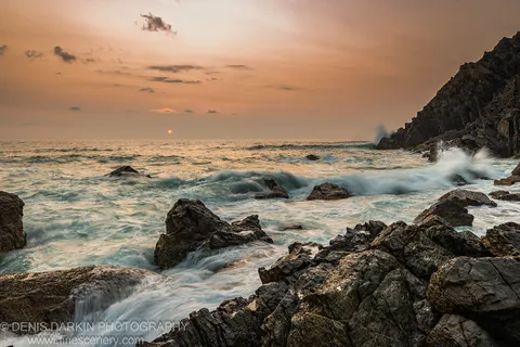 Dark jagged rocks and wave foam line the Queensland coast at dawn, with a warm amber horizon reflecting across the wet stone.