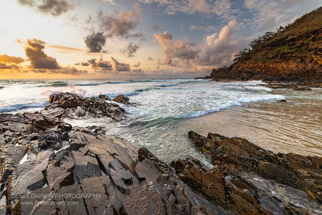 Jagged dark basalt formations at dawn on the Queensland coast, with a sandy cove sheltered by a steep cliff behind.