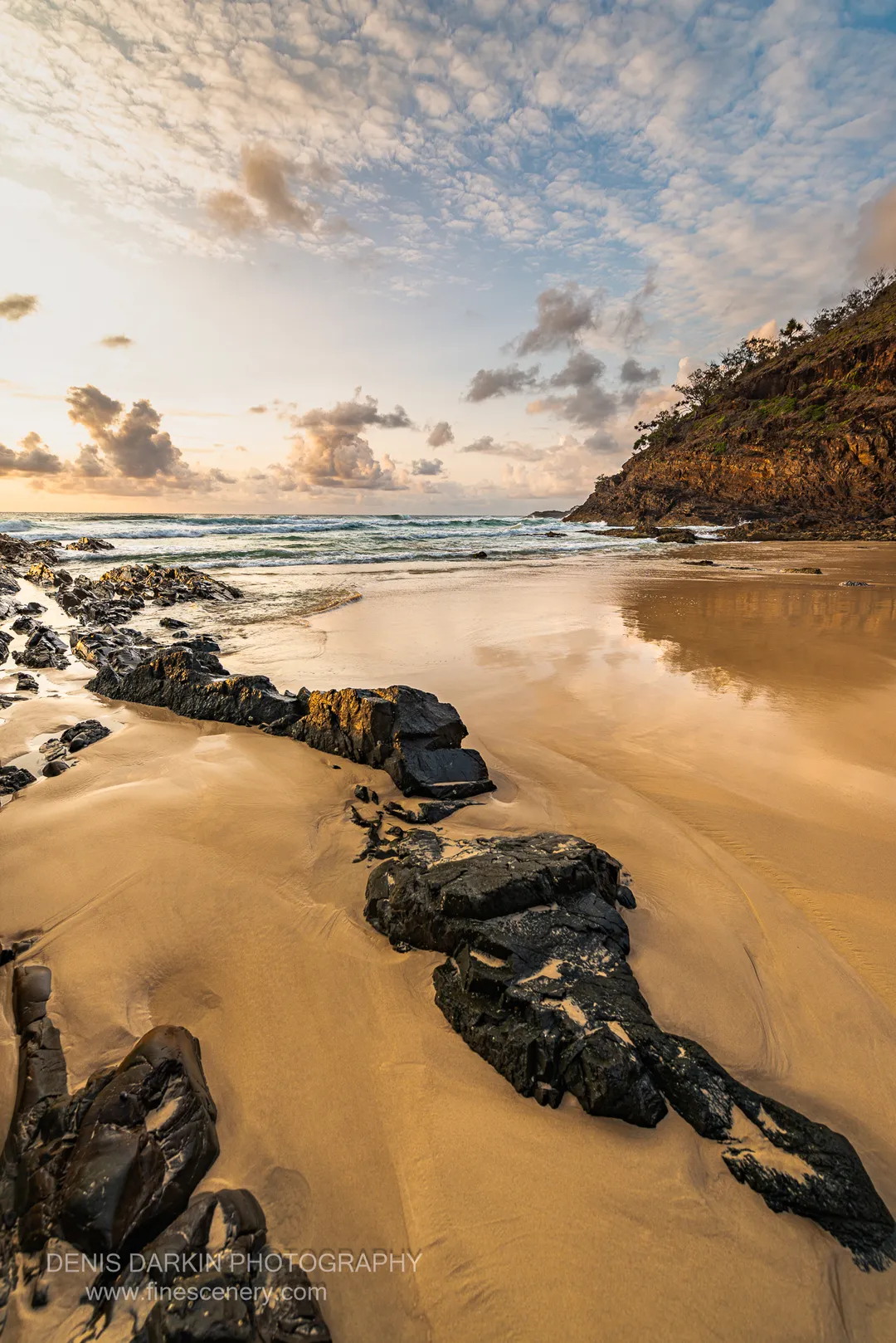Smooth wet sand on a Queensland beach reflects scattered clouds, with gentle waves and a steep cliff at the far edge.