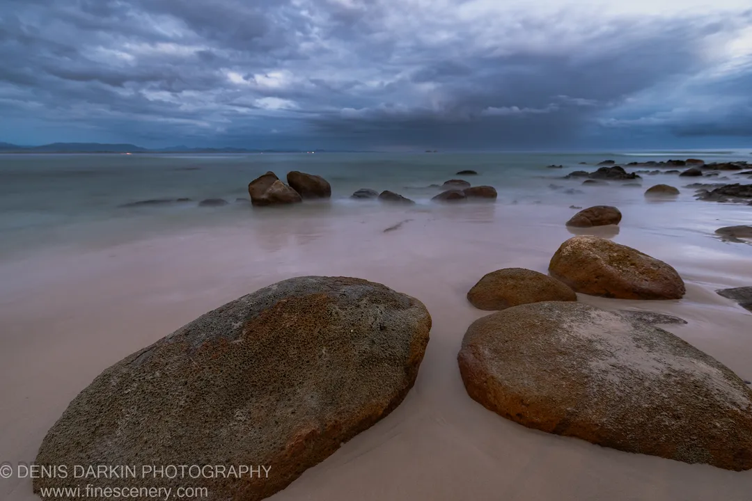 Long-exposure predawn on a Queensland beach, boulders anchoring the foreground as the glassy ocean surface reflects heavy textured cloud, January 2020.