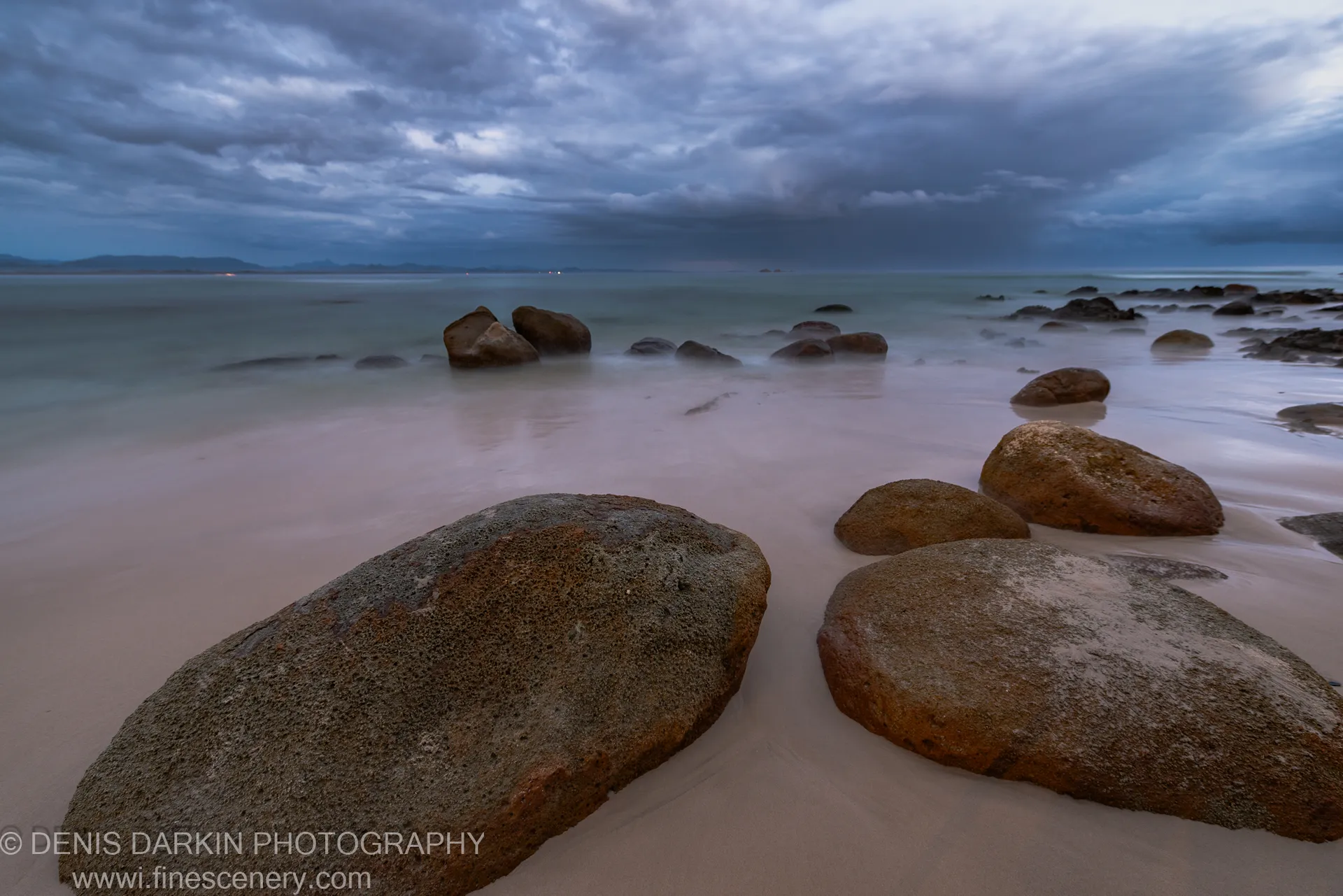 Long-exposure predawn on a Queensland beach, boulders anchoring the foreground as the glassy ocean surface reflects heavy textured cloud, January 2020.