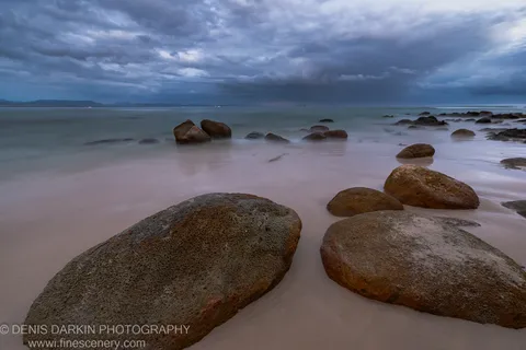 Long-exposure predawn on a Queensland beach, boulders anchoring the foreground as the glassy ocean surface reflects heavy textured cloud, January 2020.