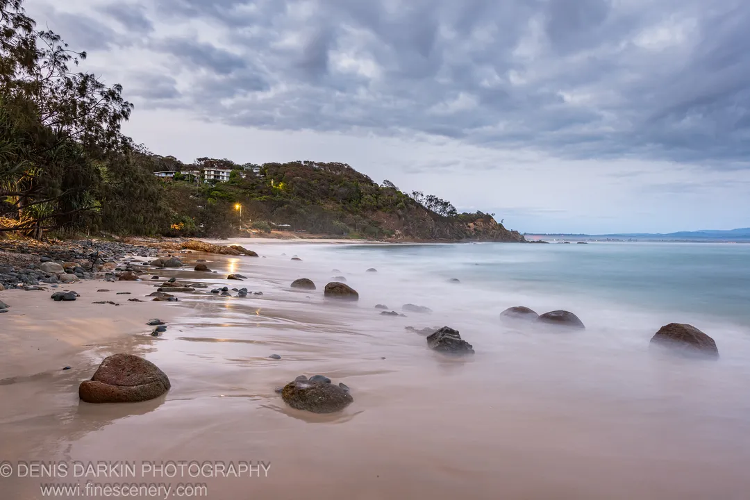 Scattered boulders on a Queensland beach at blue hour, long-exposure ocean reduced to pale mist on wet sand, a forested headland visible at the left.