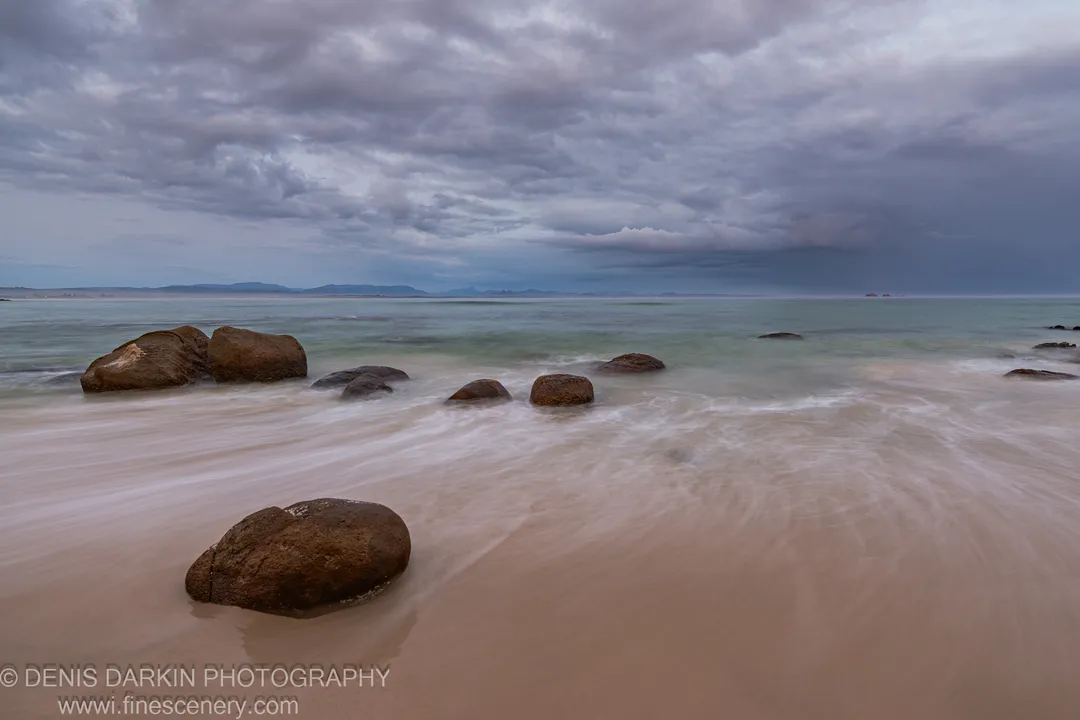 Granite boulders on a Queensland beach at predawn, stratified slate-grey clouds filling the upper frame while long-exposure water settles between the rocks.