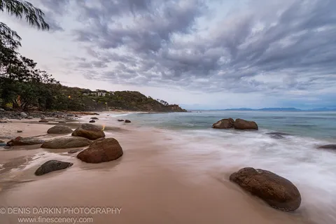 Rounded boulders on a curved Queensland beach at predawn, long-exposure water flowing between them toward a forested headland.