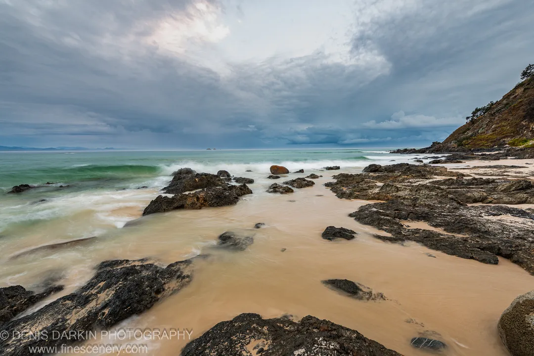 Rocky Queensland shoreline at predawn, dark coastal stones set against milky water in motion, heavy clouds and a steep hillside completing the scene.