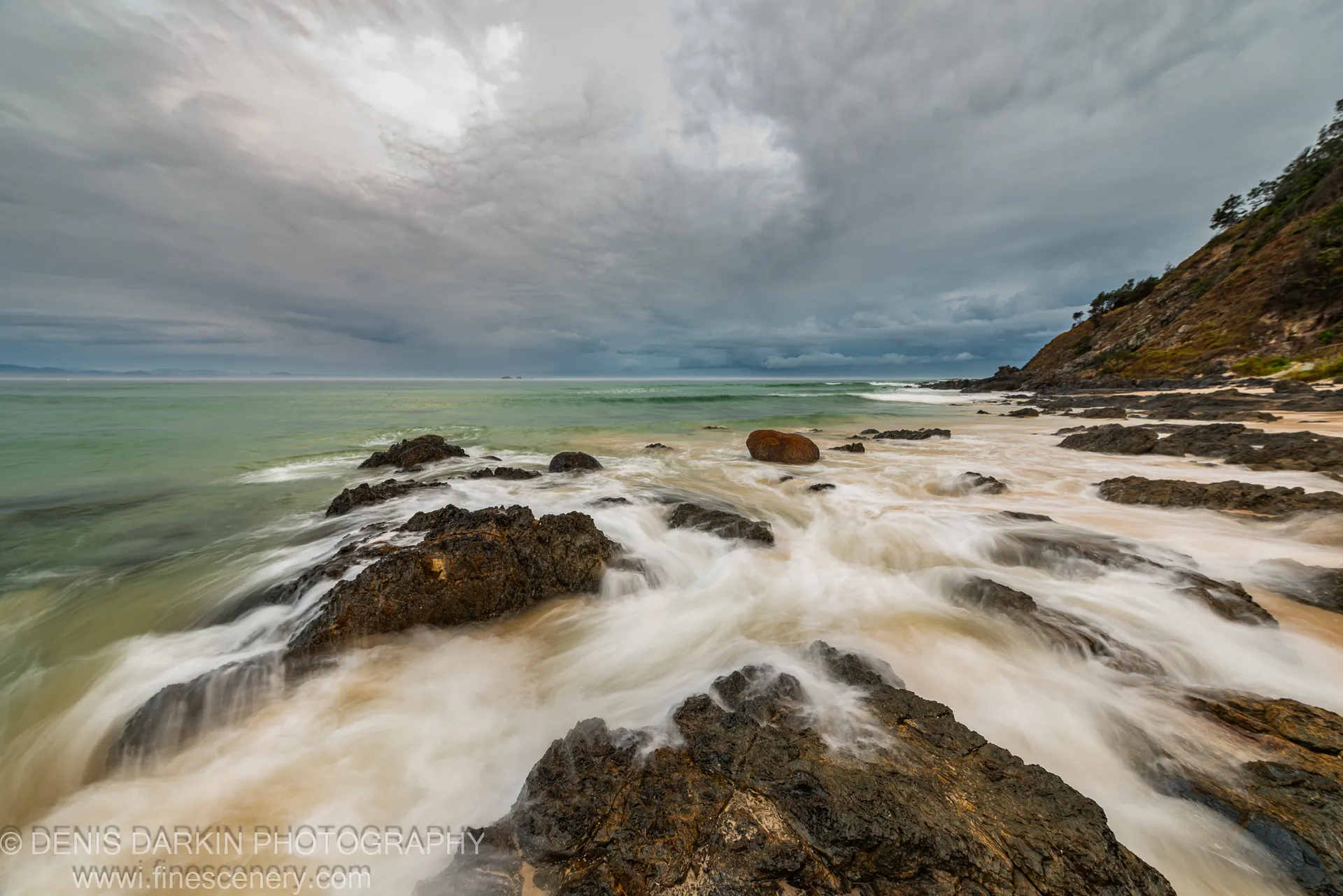Wet rubble at the base of a Queensland coastal cliff at predawn, silky water surrounding the foreground stones under a grey overcast sky.