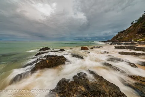 Wet rubble at the base of a Queensland coastal cliff at predawn, silky water surrounding the foreground stones under a grey overcast sky.