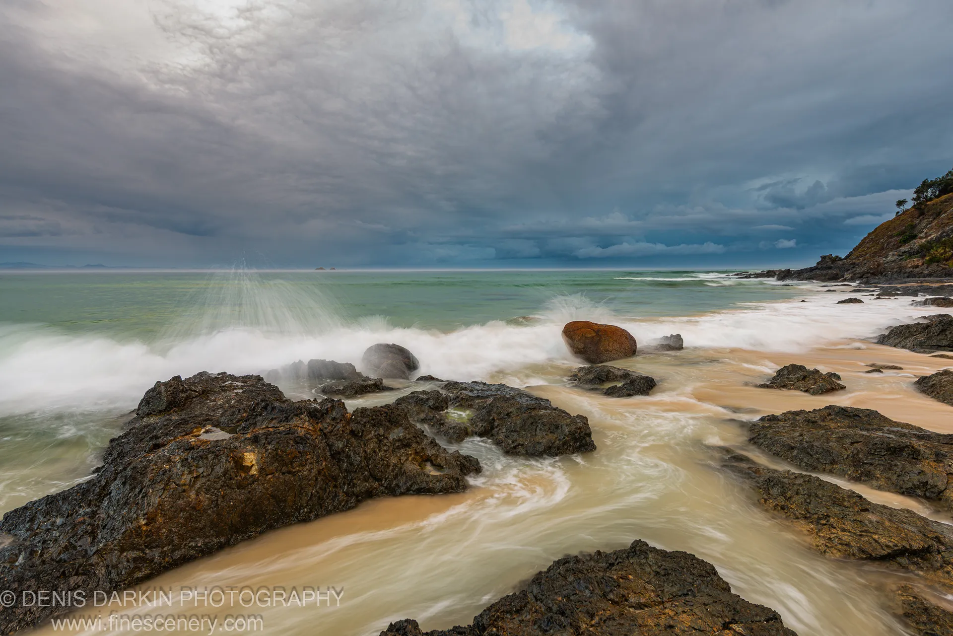 Dark wet rock formations line a Queensland coast at predawn, long-exposure waves folding into white foam under heavy cloud cover.