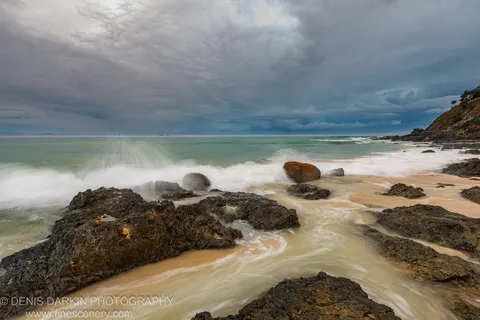 Dark wet rock formations line a Queensland coast at predawn, long-exposure waves folding into white foam under heavy cloud cover.