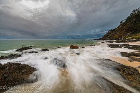 Heavy storm clouds hang above a rocky Queensland headland at predawn, with water moving through boulders toward a vegetated coastal hillside.