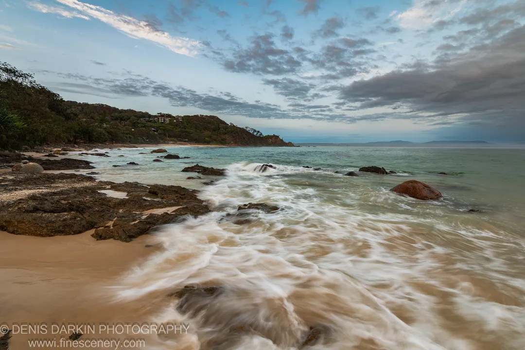 Milky tide washes over dark rock and pale sand at sunrise on the Queensland coast, grassy hillside and grey-blue sky above.