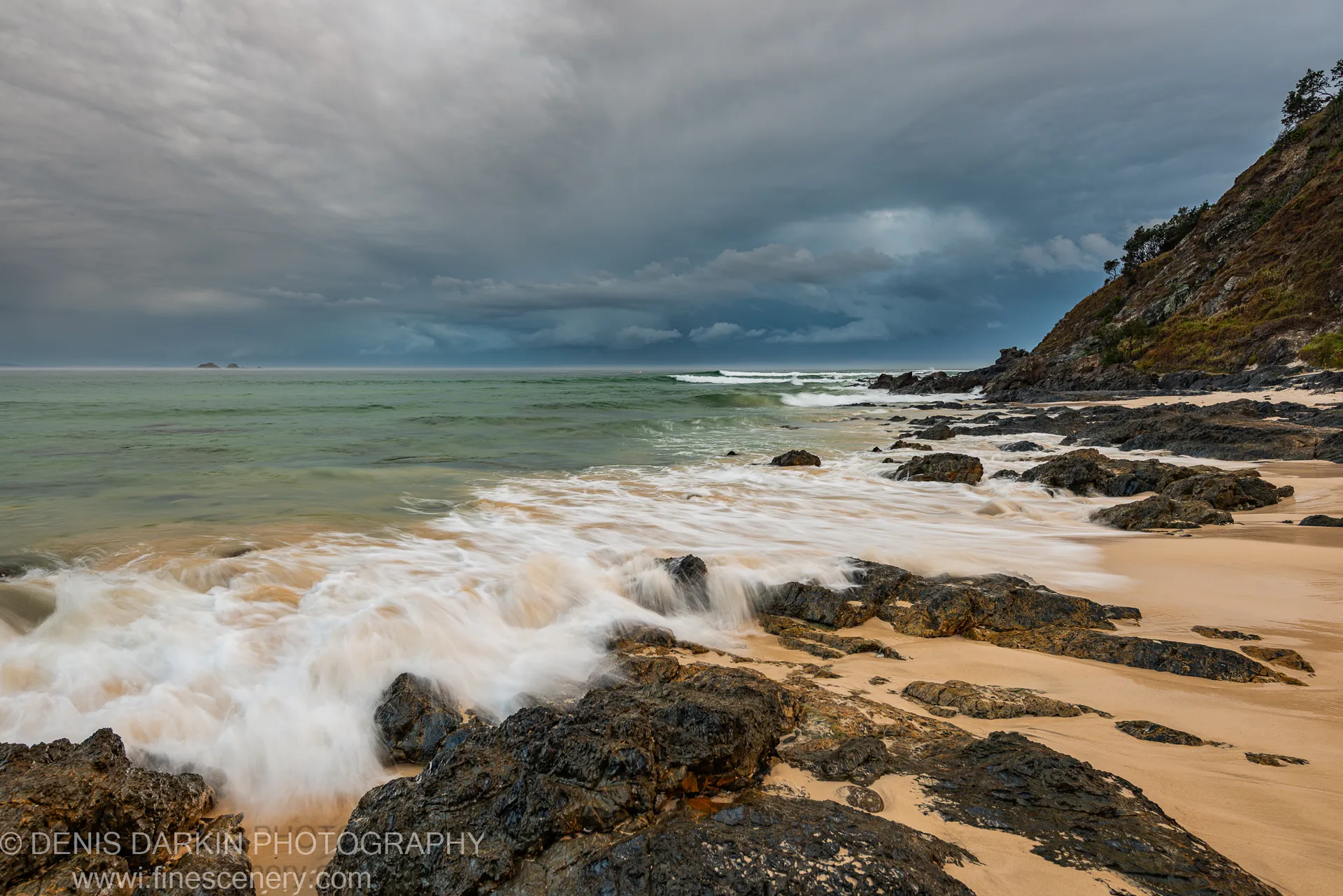 Wet basalt rocks anchor the foreground as long-exposure waves run across a sandy Queensland beach beneath heavy storm cloud.