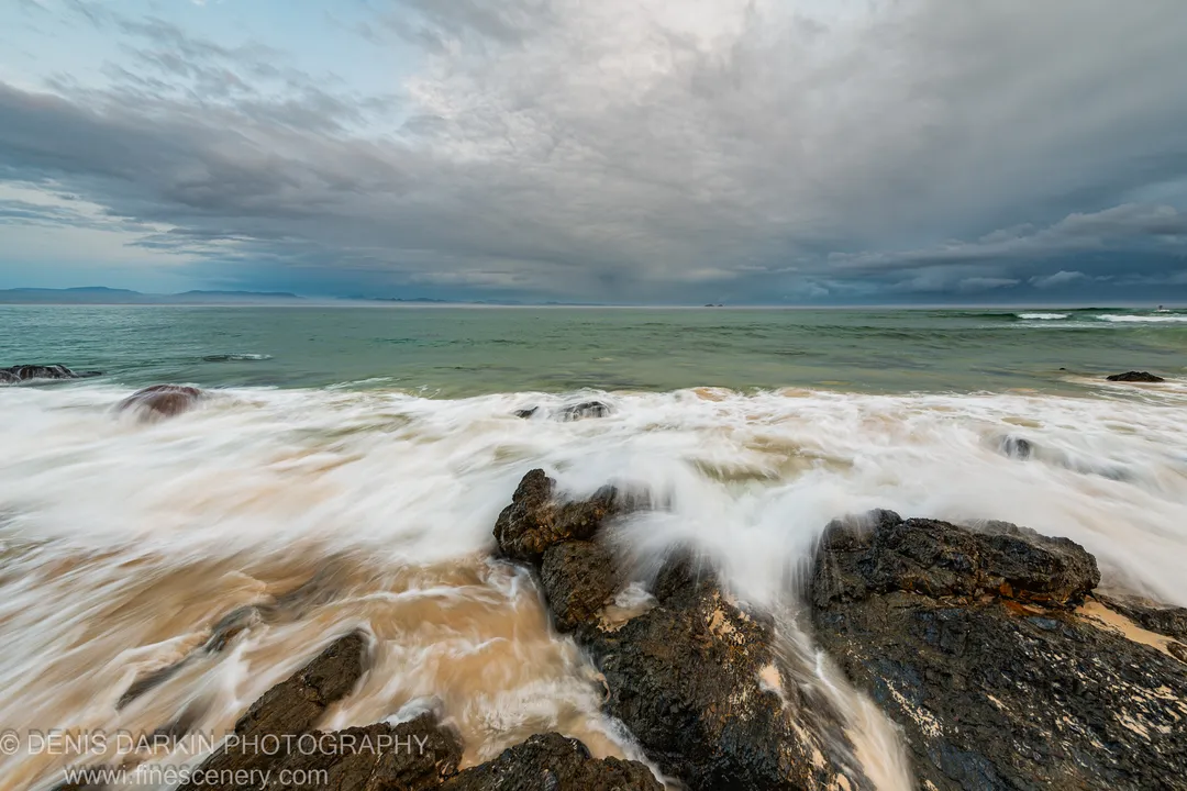 Long-exposure surf flows over dark jagged rocks on the Queensland coast under a vast overcast sky, with green sea beyond.