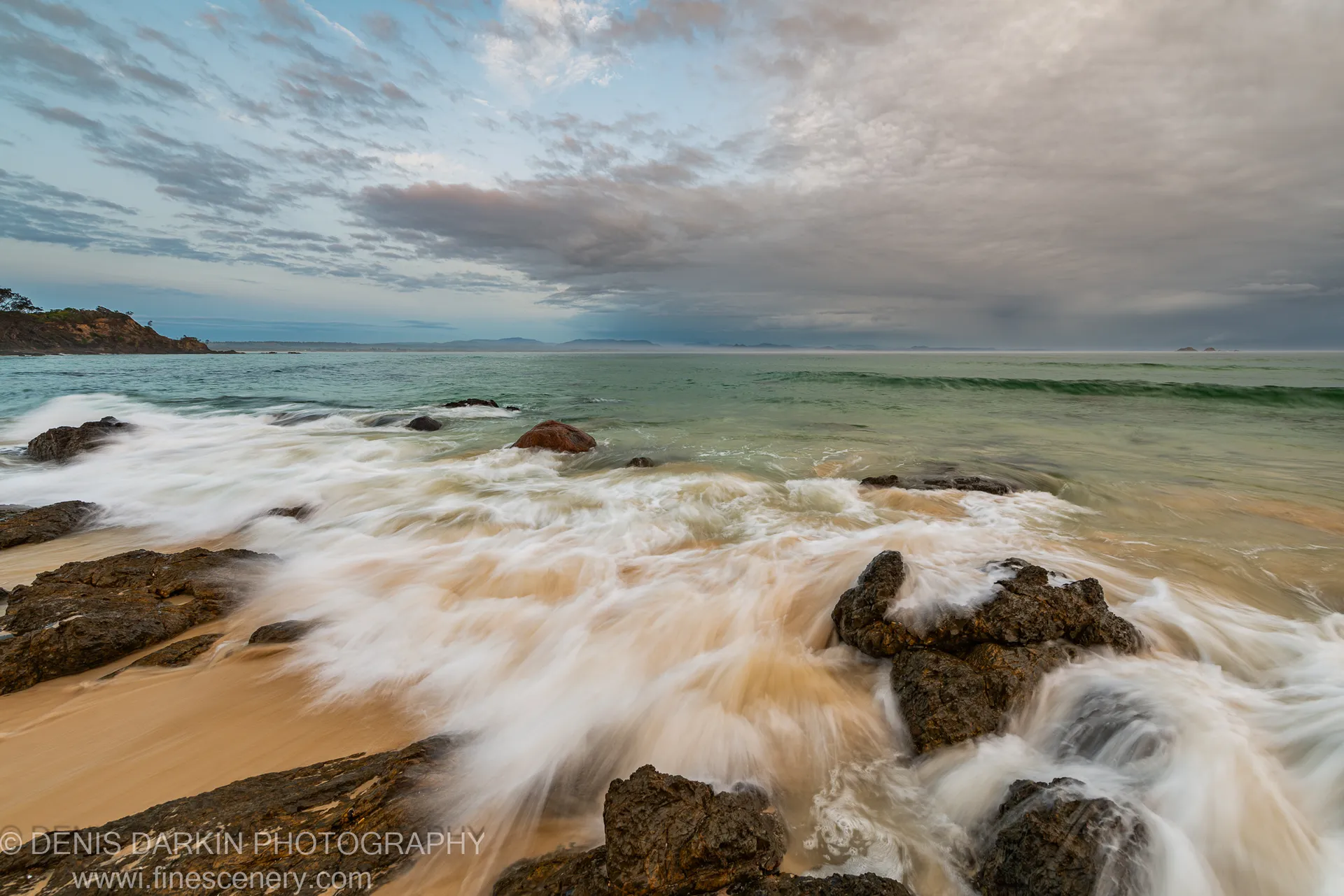 Soft milky surf flows over dark wet rocks along the Queensland coast, with a distant headland under a cloud-filled sky at dawn.
