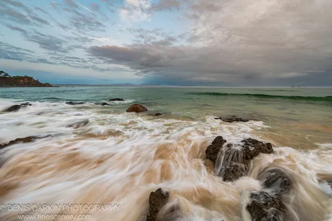 Long-exposure surf around dark coastal boulders on the Queensland coast at dawn, with a vegetated headland to the left and rain squall clouds overhead.