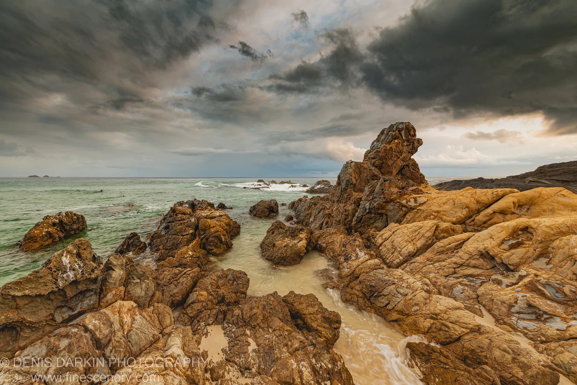 Textured sandstone in the foreground leads toward a long-exposure seawater channel on the Queensland coast, heavy storm clouds overhead at dawn.