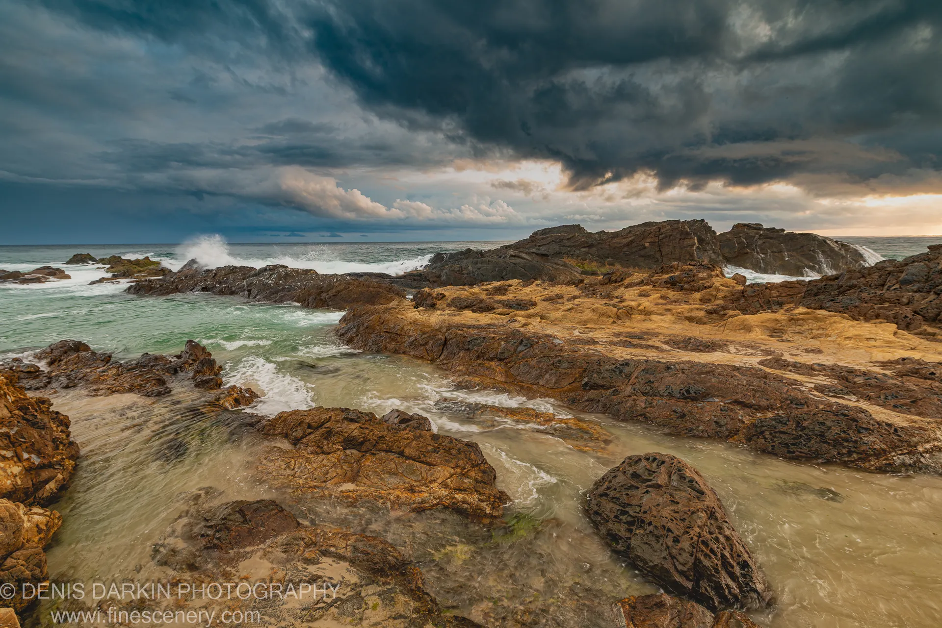 Rock pools and ochre stone formations on a Queensland headland at dawn, storm clouds above with warm light breaking through from the right.