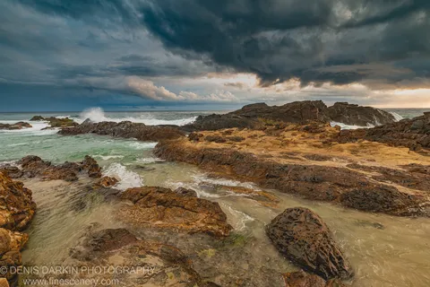 Rock pools and ochre stone formations on a Queensland headland at dawn, storm clouds above with warm light breaking through from the right.