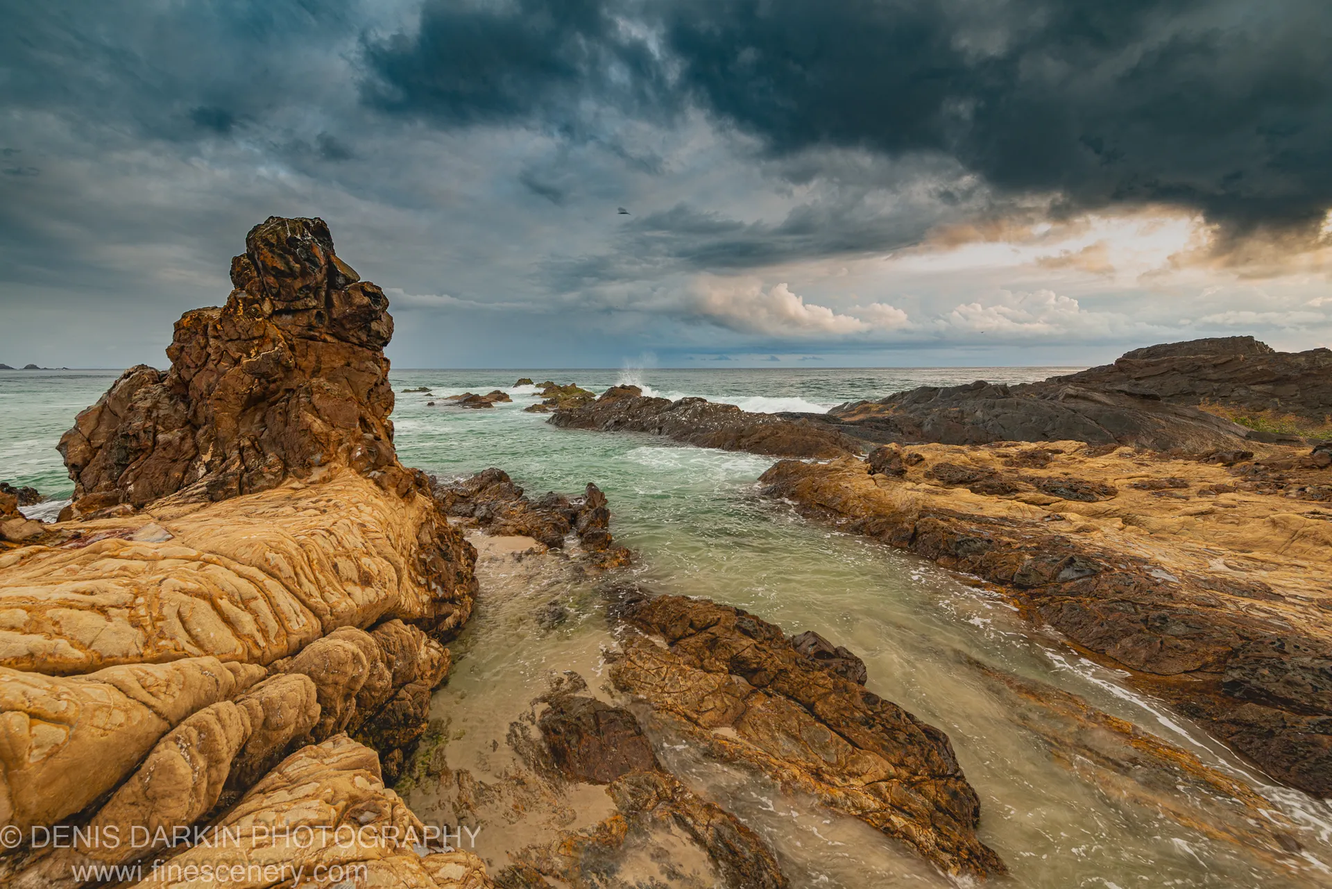 Deeply grooved sandstone boulders beside a calm tidal channel on the Queensland coast at dawn, heavy clouds and white foam in the background.