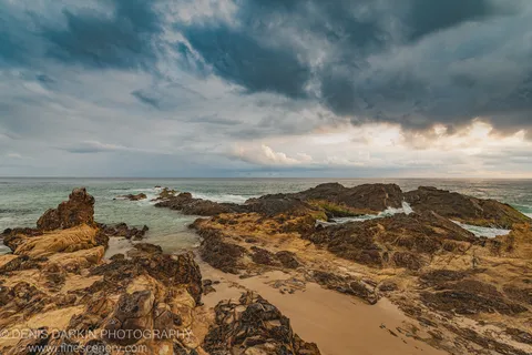 Warm morning light breaks through slate storm clouds over a rocky Queensland cove, lighting the foreground rocks and turquoise water.